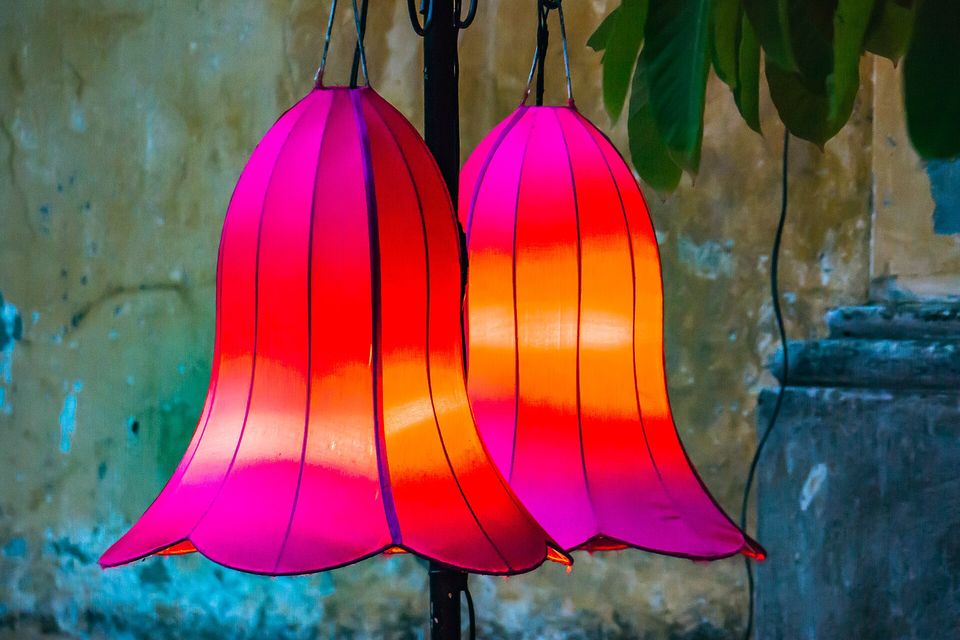 Two bell-shaped silk lanterns glowing magenta and orange against a peeling teal and ochre wall, green leaves at the top right