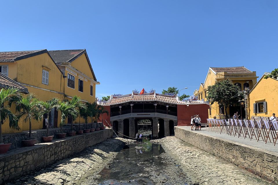 A tiled covered bridge between yellow houses at the end of a stone-lined canal drained to a muddy bed, under a bright blue sky