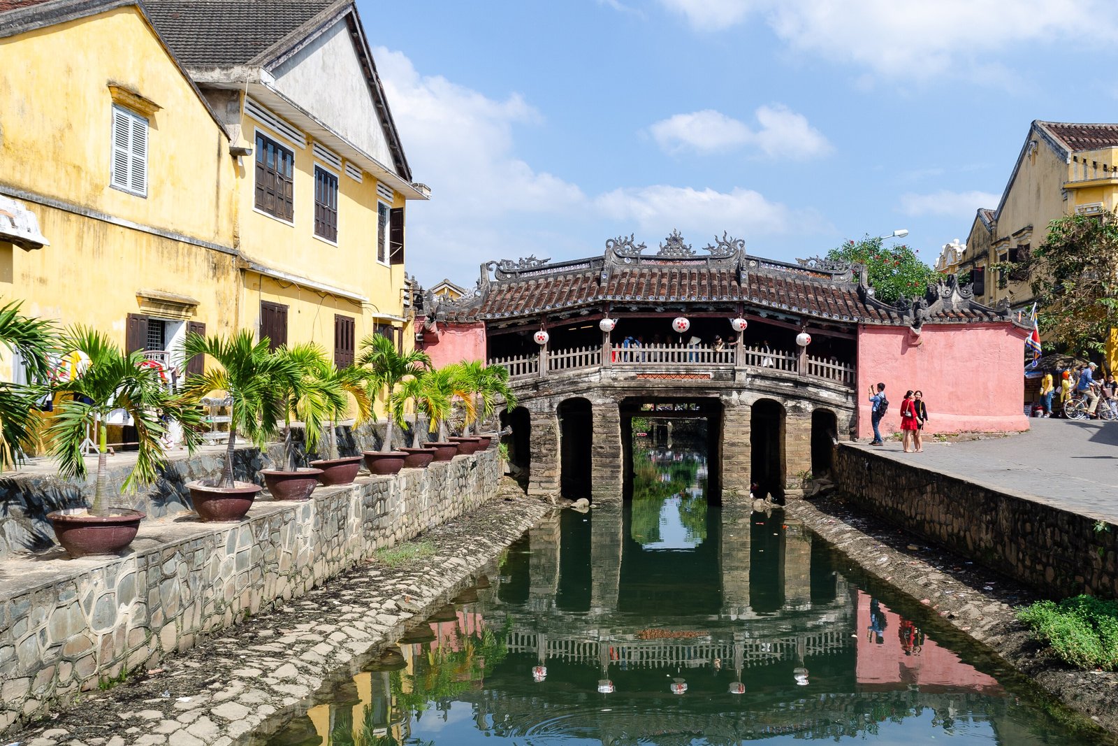 A tiled covered bridge with a small temple on top, reflected in a still canal between yellow-ochre houses, under a blue sky with white cloud