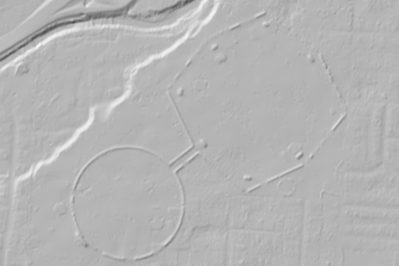 A shaded relief view from above showing a large circular earthwork joined by a short causeway to an eight-sided enclosure, their low walls picked out by raking light