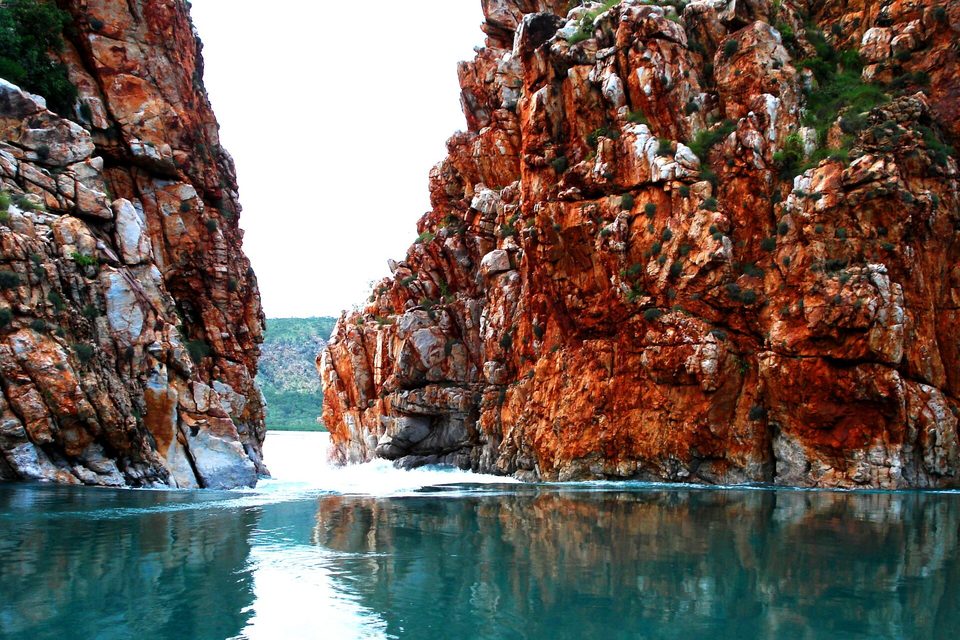 Rust-red cliffs on both sides of a narrow gap, white water pouring through it onto a smooth green-blue pool, a green hill seen through the gap