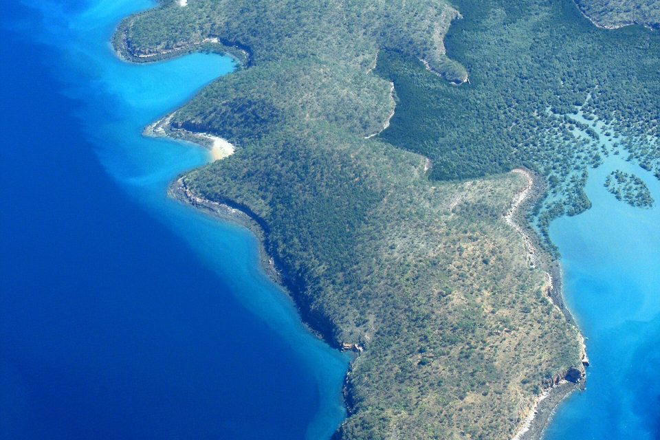 A wooded coastline seen from the air, green ridges cut by pale inlets, mangrove flats and turquoise shallows along the shore, deep blue water at the left