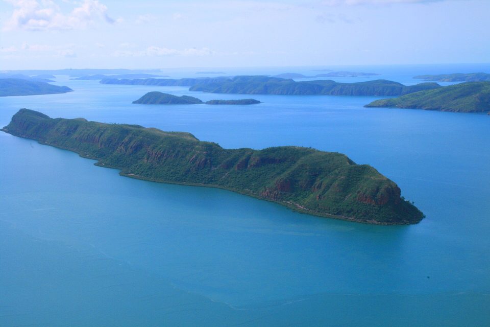 A long green-topped island with red cliffs in a pale blue sea, more low islands strung out to a hazy horizon