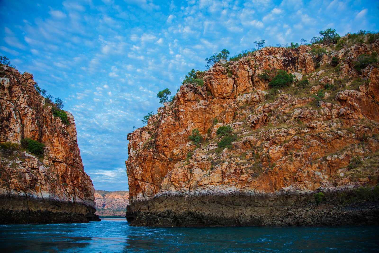 Garaan-ngaddim (Horizontal Falls), Dambimangari Country: two red-orange rock walls with a narrow gap between them over still blue-green water, a far range seen through the opening, under a sky of small clouds