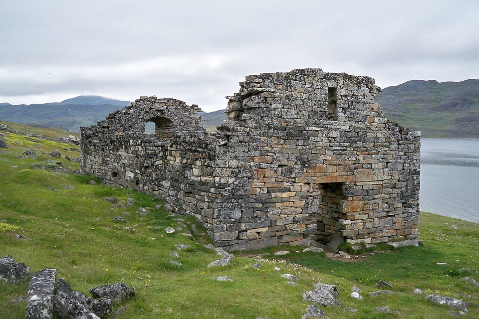 A long, roofless stone church with an arched window in its gable, on green grass beside grey water