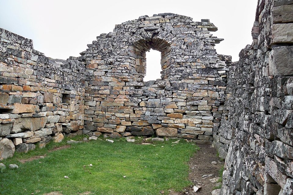 A stone gable wall with a tall arched opening, seen from inside a grassy, roofless nave