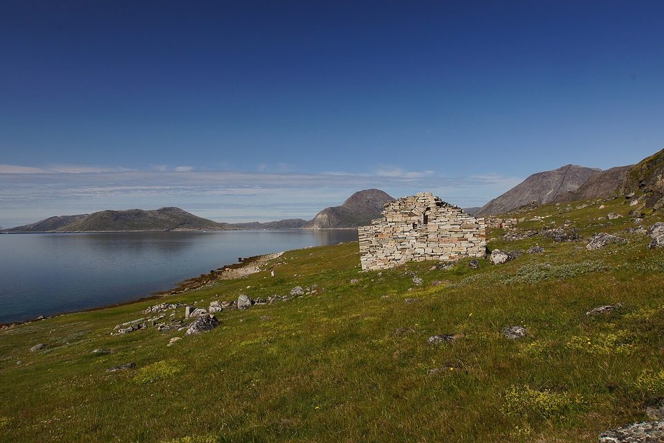 A stone gable stands alone on a green slope above a calm fjord with low mountains beyond