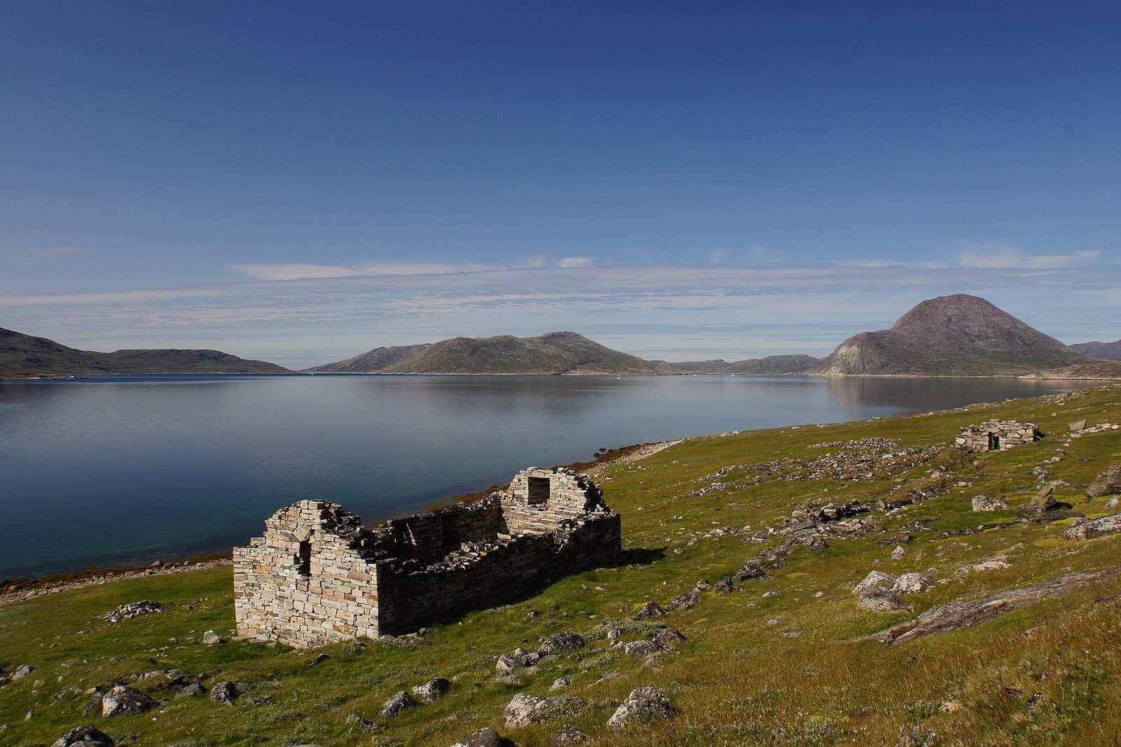 A roofless granite church stands low on a green shore beside a calm fjord, with a cone-shaped peak beyond