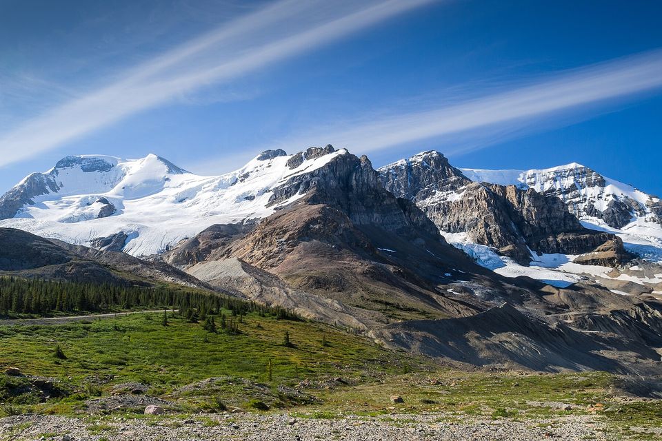 Snow-capped peaks and hanging glaciers above bare grey moraine and gravel flats, a strip of green meadow and the road in the foreground