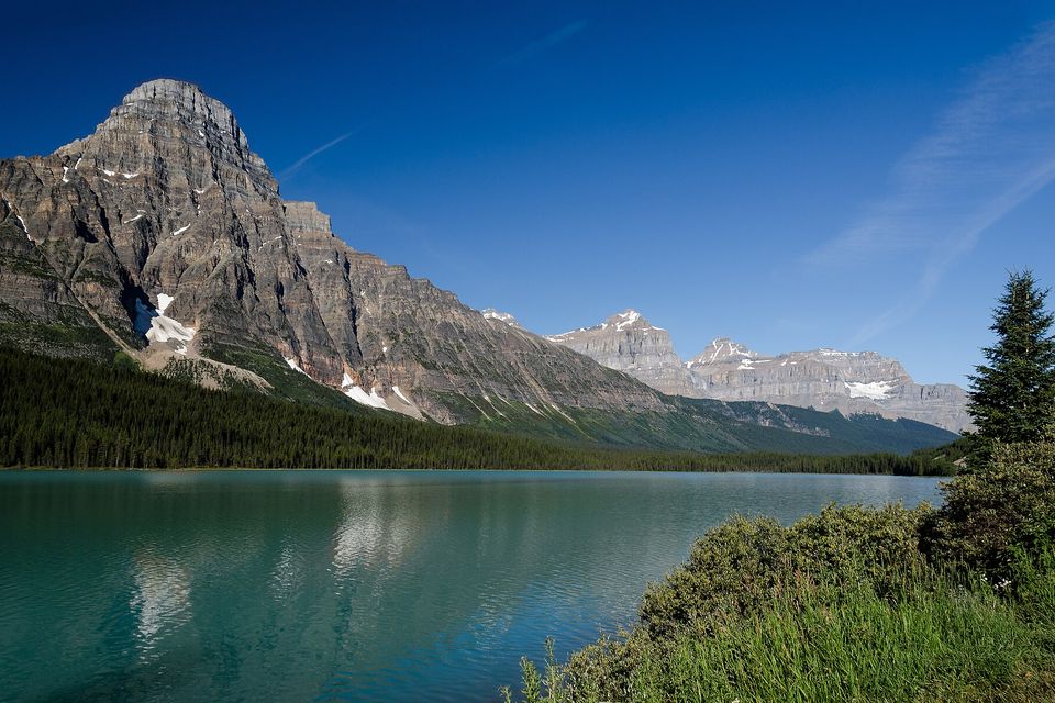 A tall pyramid of layered grey rock rising above a calm turquoise lake, dark conifers along the shore and grass in the foreground