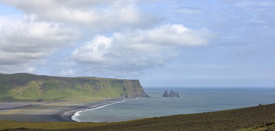 A long black-sand beach under a green headland, rock stacks standing in the sea off its end beneath a cloudy sky