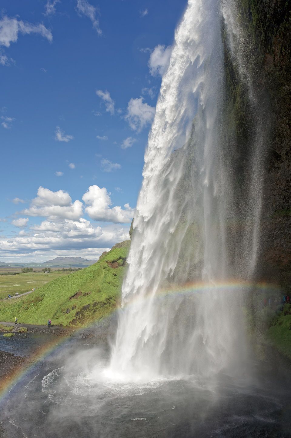 A tall waterfall seen from beside it with a rainbow across its spray and green meadow below a blue sky