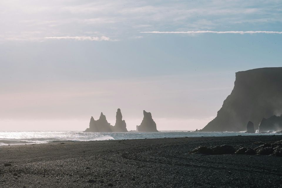 Dark rock stacks standing in the sea beside a high cliff, a black-sand beach in the foreground, in pale hazy light