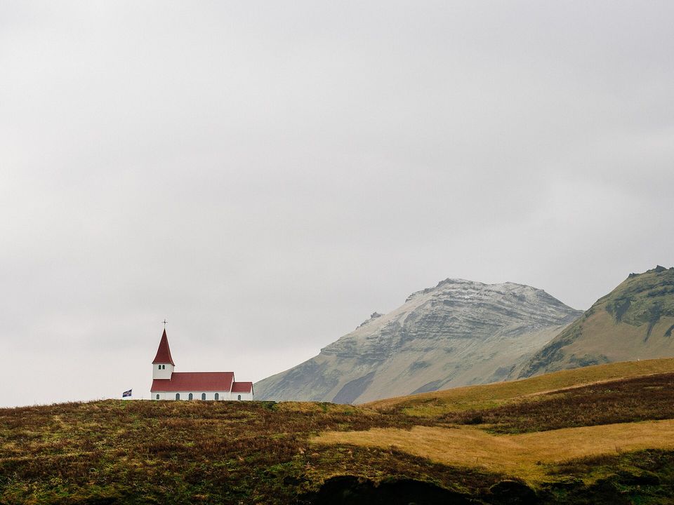 A small white church with a red roof on a hilltop, brown moorland and pale mountains behind under an overcast sky