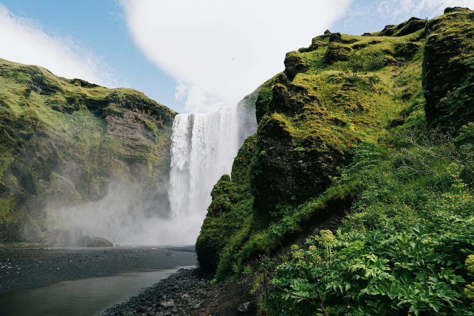 A tall waterfall dropping between steep moss-green cliffs, mist rising at its foot under a bright sky