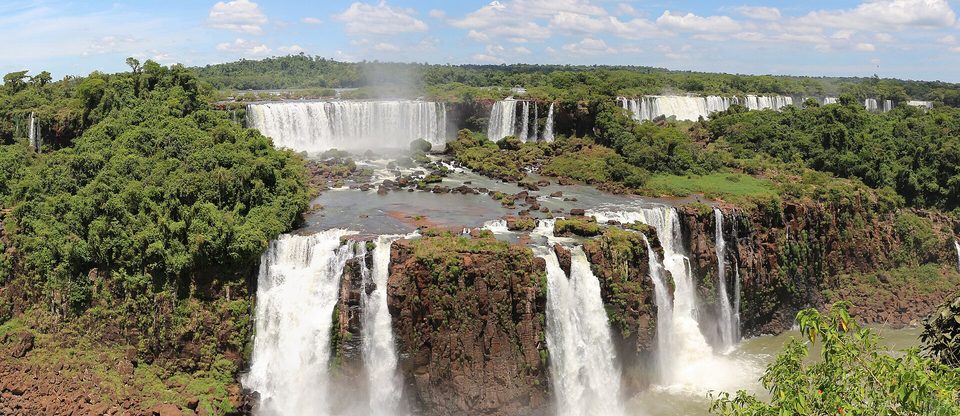 A wide view of many waterfalls tumbling over a broad cliff edge, with forest in the foreground and a blue sky with scattered cloud