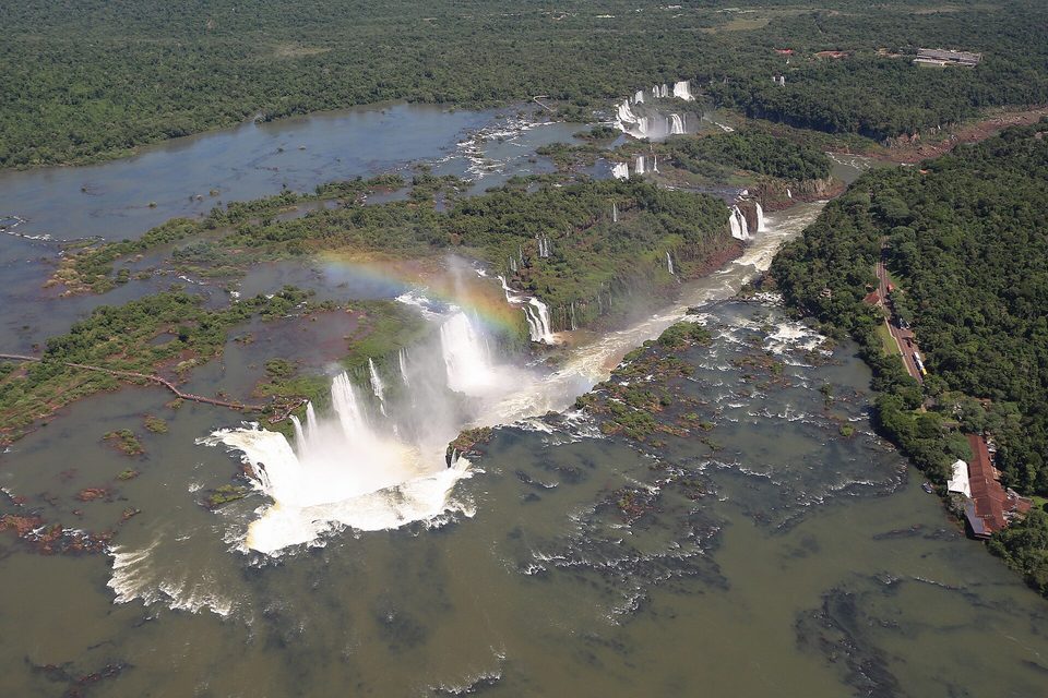 An aerial view of a horseshoe of white water plunging into a gorge with a rainbow above it, a walkway crossing the wide brown river at left