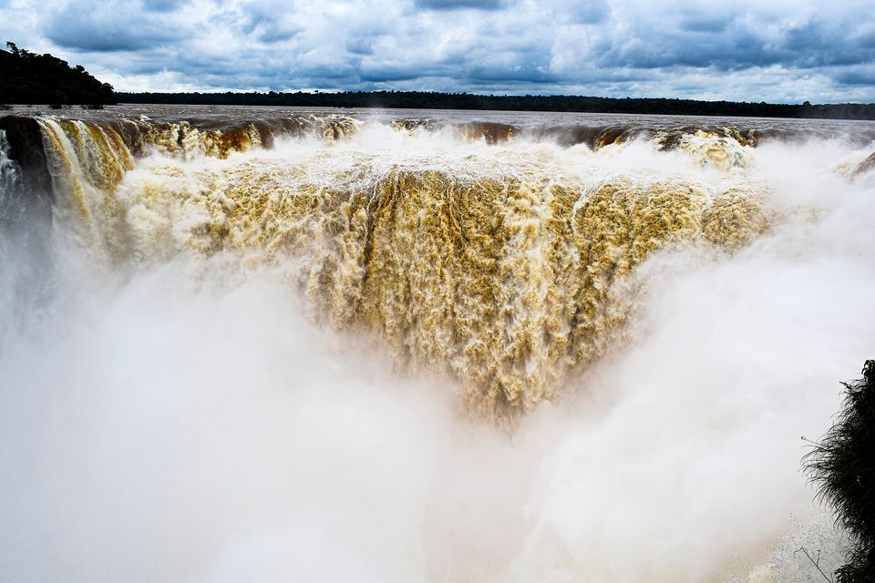 A wall of brown and white water curving into a cloud of spray, a dark forest line and stormy blue sky above