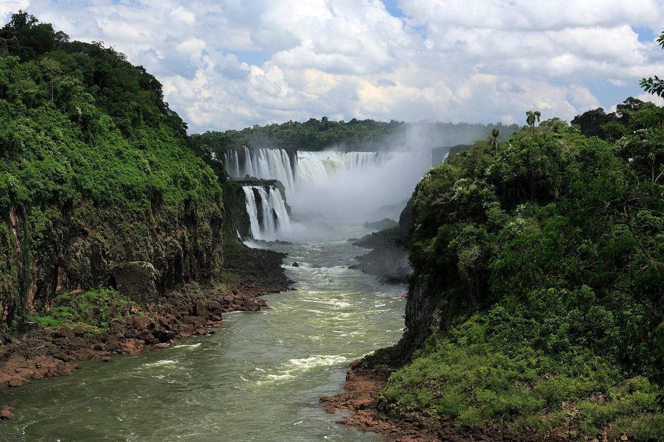 A green forested gorge with a brown river running away from a distant curtain of spray and waterfalls, cloud above