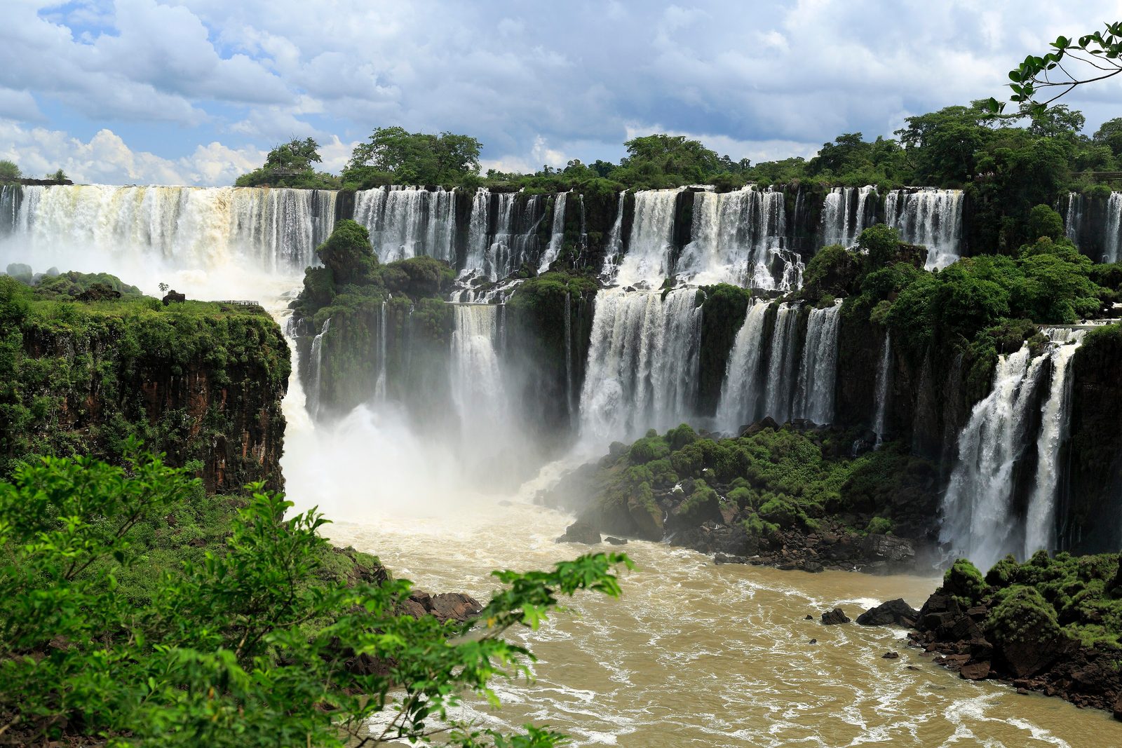 A long row of white waterfalls dropping over a dark forested cliff into a brown river, with green trees below and broken cloud above