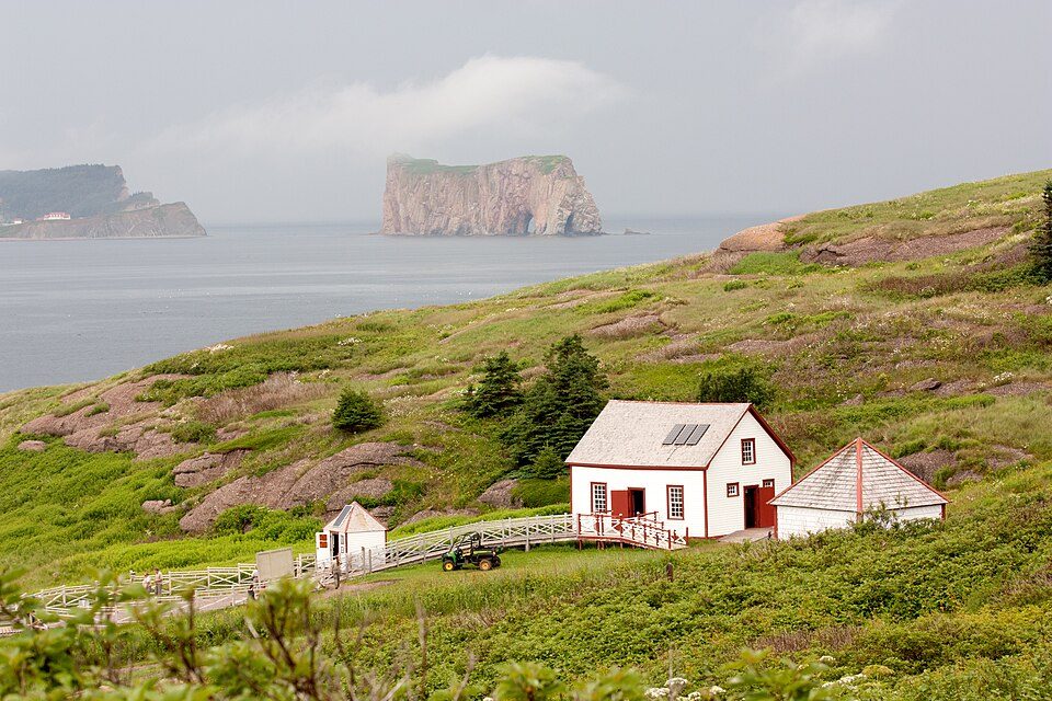 A white clapboard house with red trim sits on a green slope above the sea, a sheer rock island with an arch beyond in mist