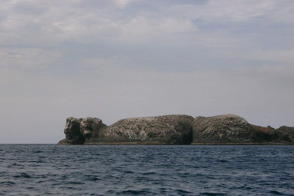 A long, low basalt islet with steep dark sides and a pale, streaked top sits on a grey-blue sea
