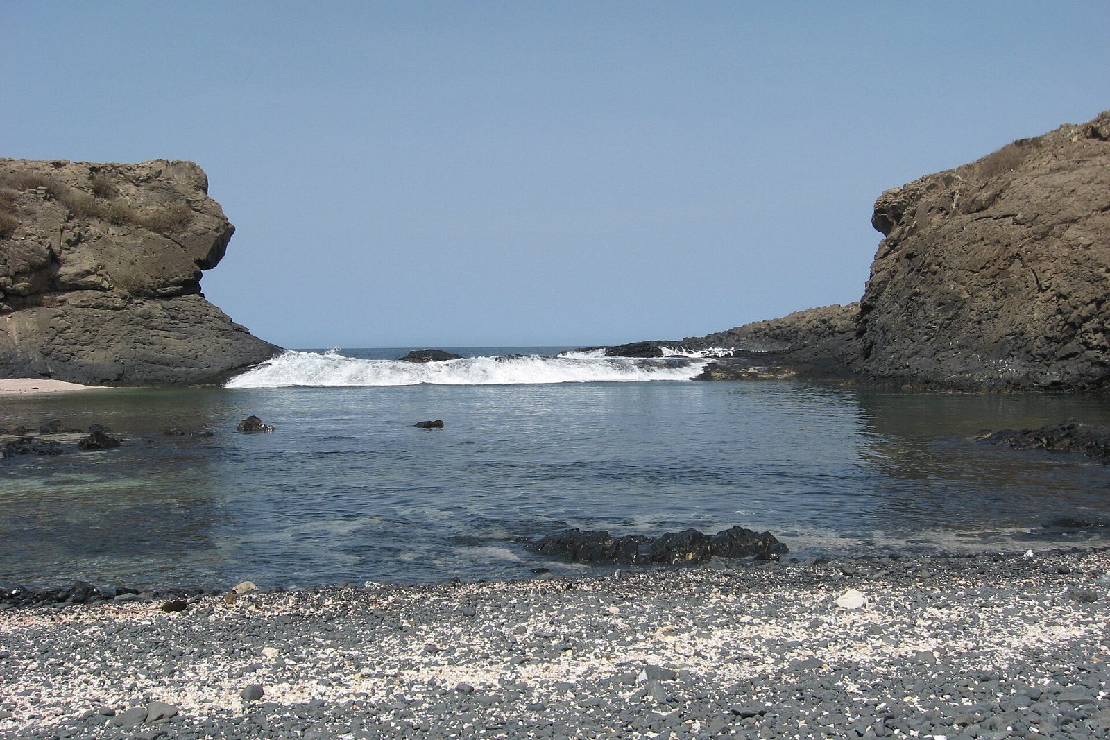 A pebble beach flecked with white shell leads to a calm inlet where surf breaks through a gap between dark volcanic cliffs