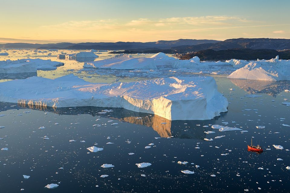 Icebergs of many sizes on flat blue water in low golden light, one small red boat in the middle distance