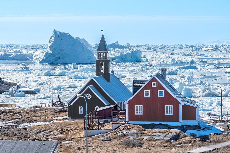 A dark wooden church and a red wooden house in the foreground with ice-choked water and large icebergs behind
