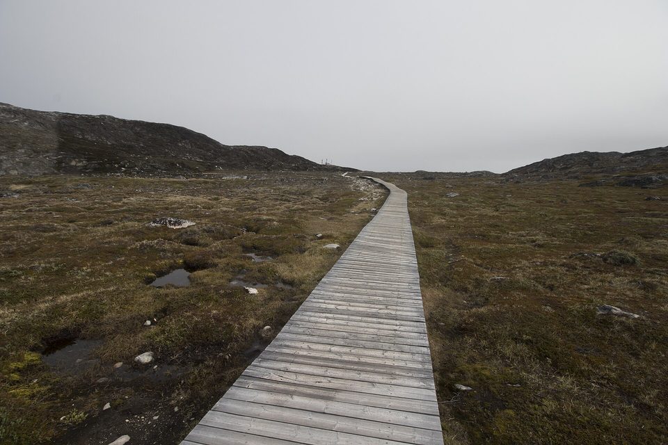 A wooden boardwalk running across brown tundra toward low hills under a grey sky