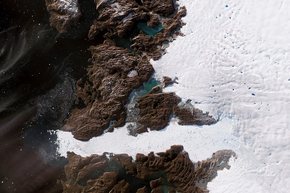 A satellite view of a white glacier tongue flowing between dark brown rock into a fjord