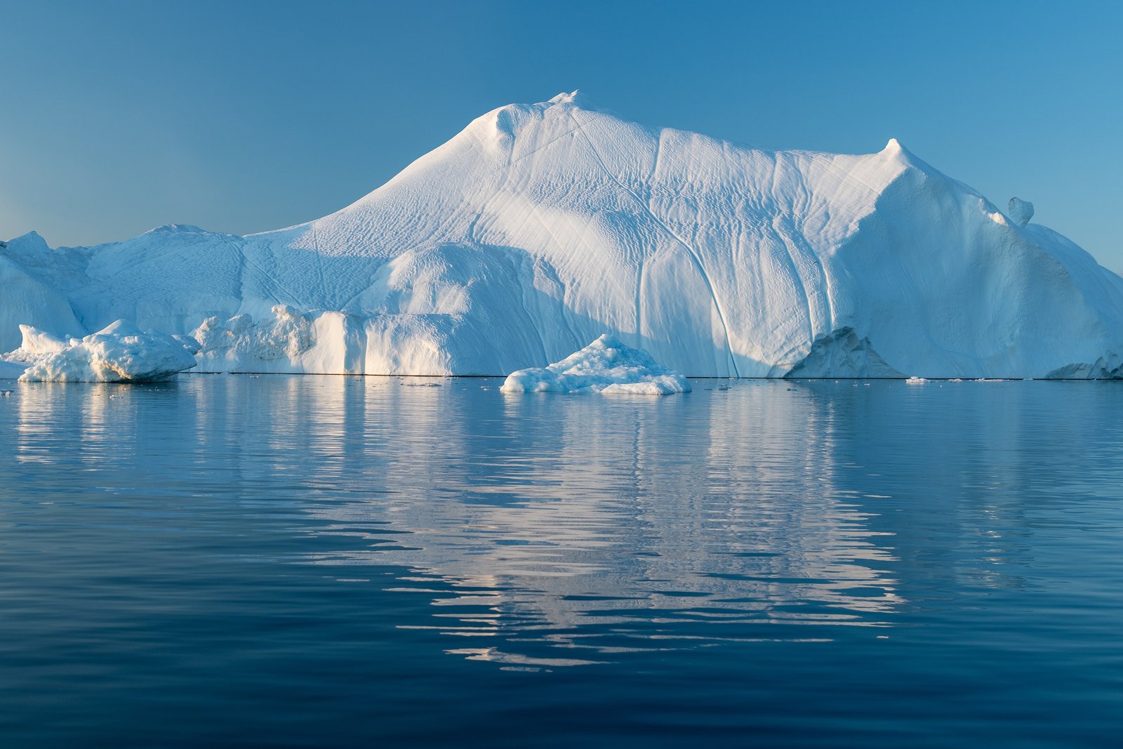 A large white iceberg with sculpted sides floating in flat blue water that mirrors it, under a clear sky