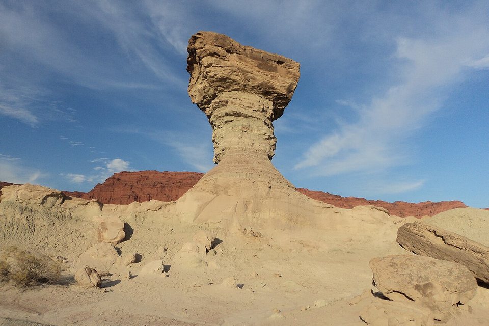 A capped column of sandstone against a blue sky with red cliffs low behind