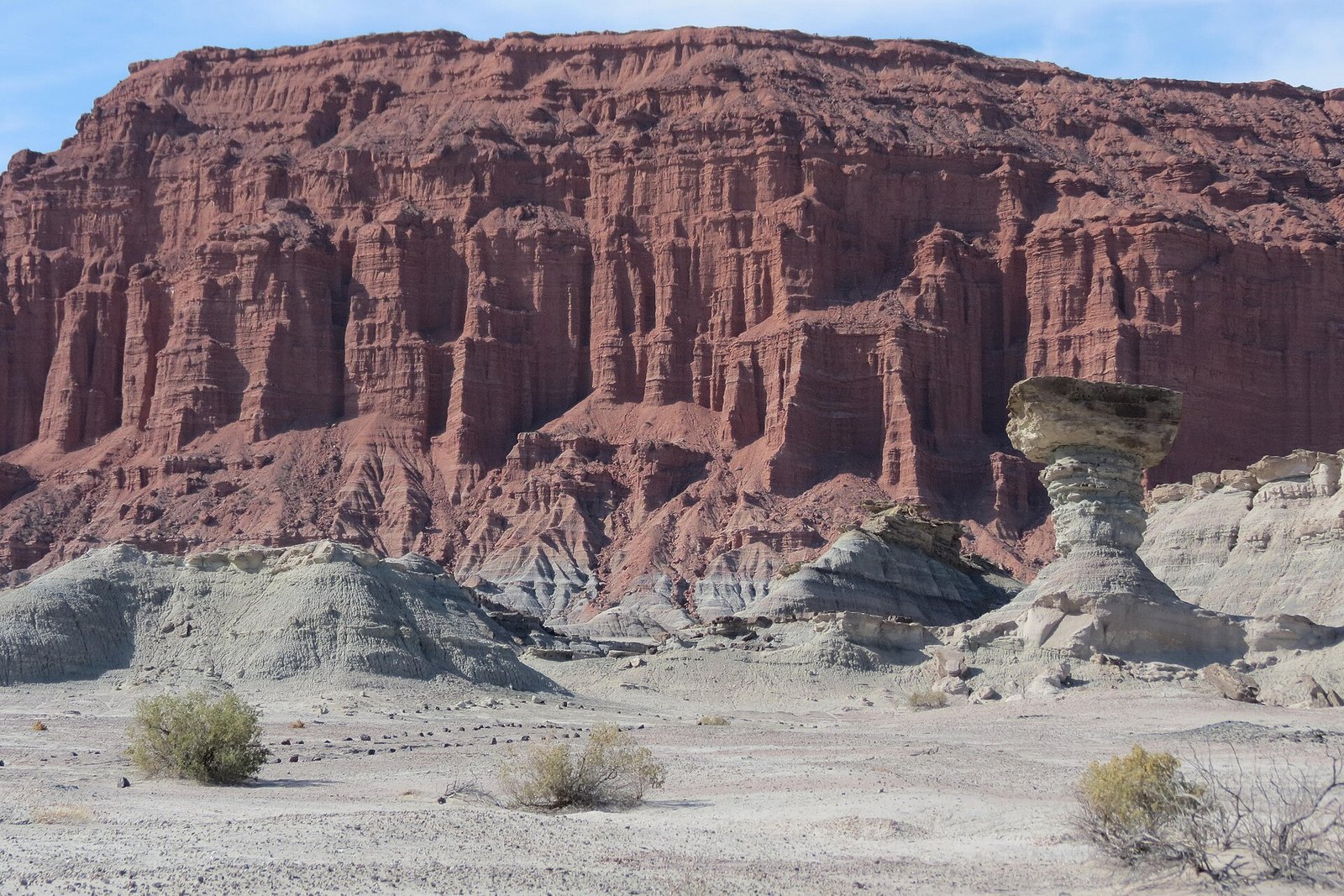 A pale mushroom-shaped rock before towering rust-red cliffs over grey-white badland