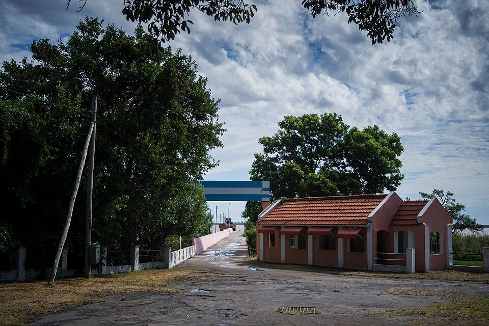 A puddled road leads past a pink tiled-roof building toward a pier under a blue-and-white arch, with trees and a cloudy sky