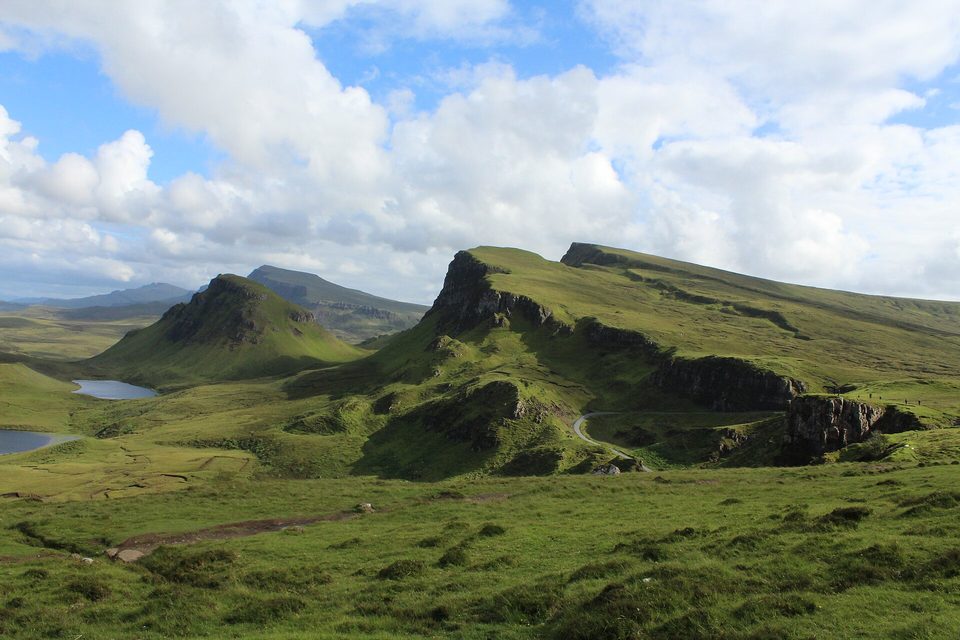 Green, flat-topped hills and crags rise above a moor dotted with small lochs, a narrow road winding between them, under big white clouds
