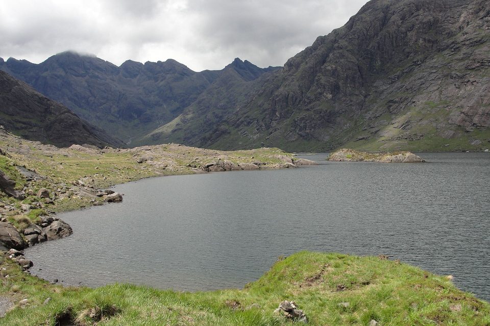 A dark loch with a small rocky islet lies in a bowl of grey, jagged mountains, a grassy bank in the foreground under low cloud