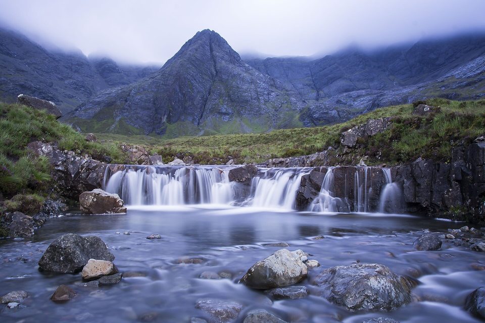 A clear stream tumbles over a wide rock step into a pool among boulders, with a steep, cloud-capped peak behind