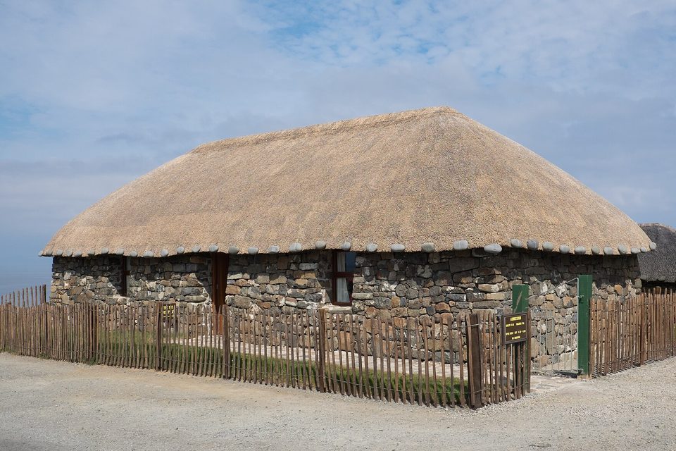 A low stone cottage with a thick, rounded thatched roof stands behind a wooden fence under a blue sky