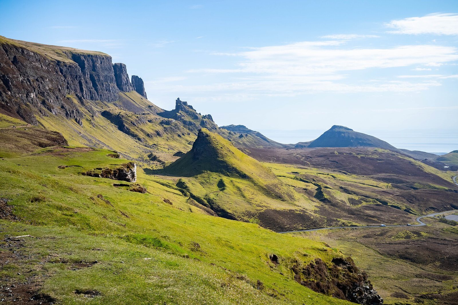 A dark escarpment runs in from the left above bright green slopes, a row of rock towers mid-frame and a sharp grassy cone in front, the moor and the sea beyond under a high blue sky