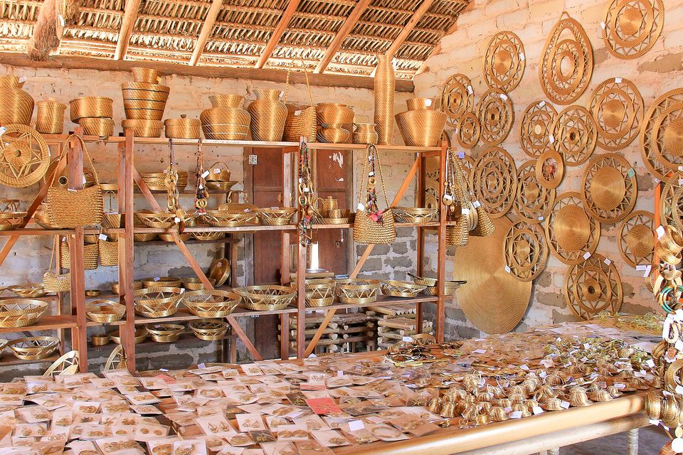 A craft shop with shelves of golden-grass baskets and bowls, round woven wall pieces on a brick wall and small goods laid out on a table below