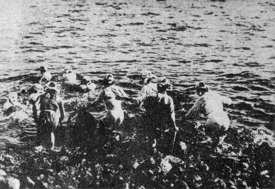 A black-and-white photograph of a row of women in light cotton clothing wading from a rocky shore into choppy water