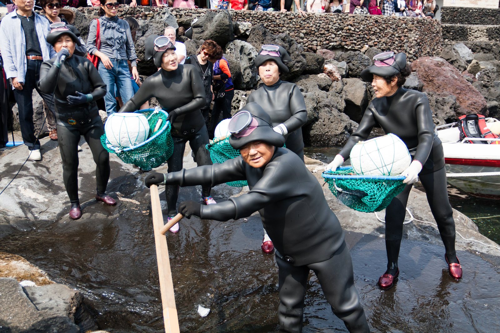 Five women in black wetsuits and hoods stepping over wet volcanic rock at the shoreline, carrying green mesh catch nets slung under round white floats