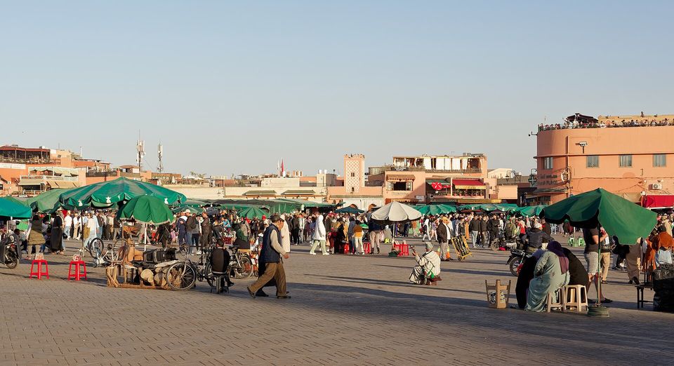 A wide paved square in warm low sun, green umbrellas over stalls along the far side and pink buildings against a pale sky