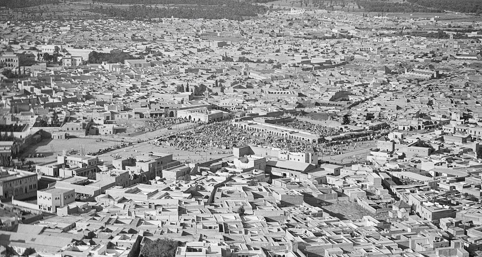 A black-and-white aerial view of a dense city of flat-roofed white houses, with a wide open square packed with a dark crowd at the centre