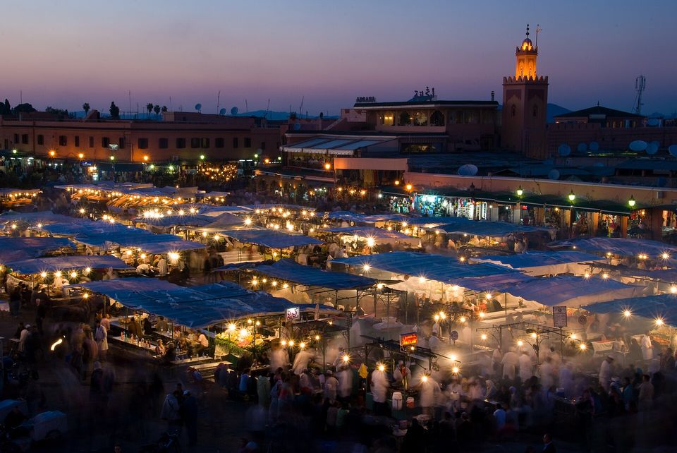 Rows of blue-roofed food stalls hung with warm lights in a dusk square, blurred figures moving beneath, and a lit minaret against a purple sky