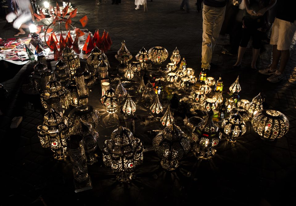 Dozens of pierced-metal lanterns glowing on dark paving, with a stand of small red Moroccan flags and the legs of passers-by behind
