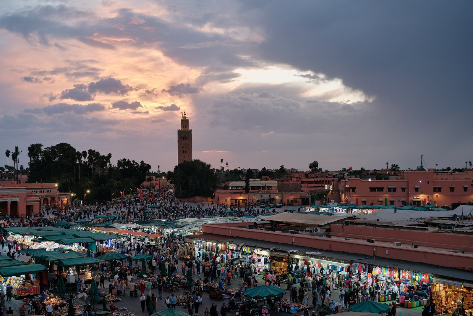 A crowded market square at dusk under a stormy pink and grey sky, green stall awnings below, low pink buildings and a tall square minaret in the distance
