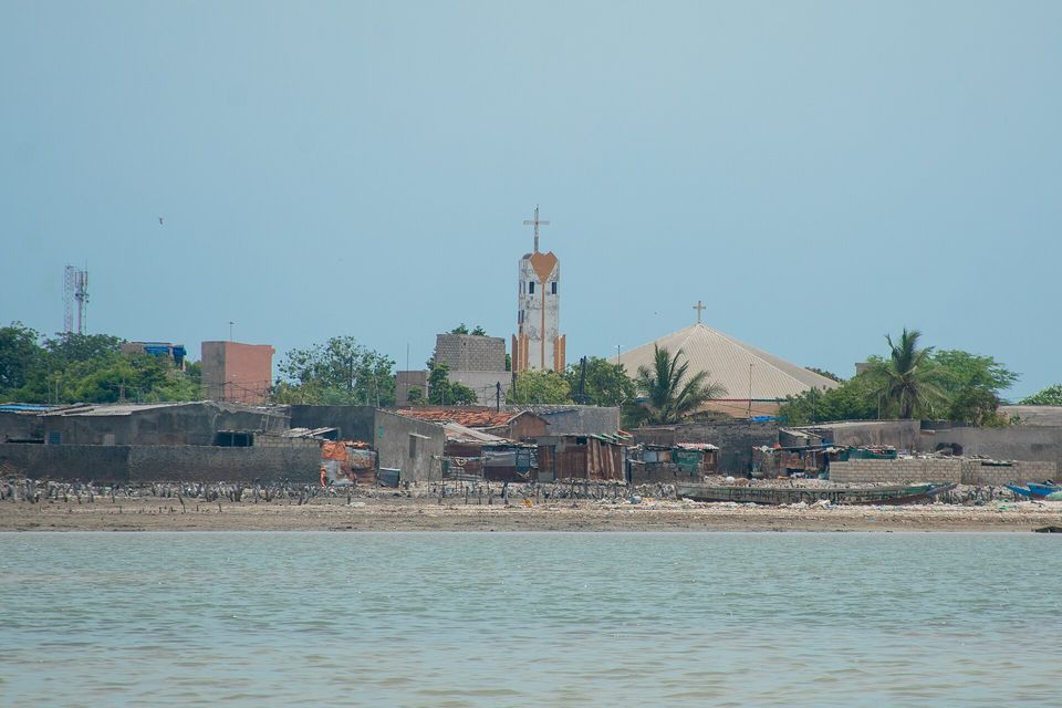 Low houses and walls line a pale shell beach, a white church tower with a cross rising behind and a second church roof at right, under a hazy blue sky