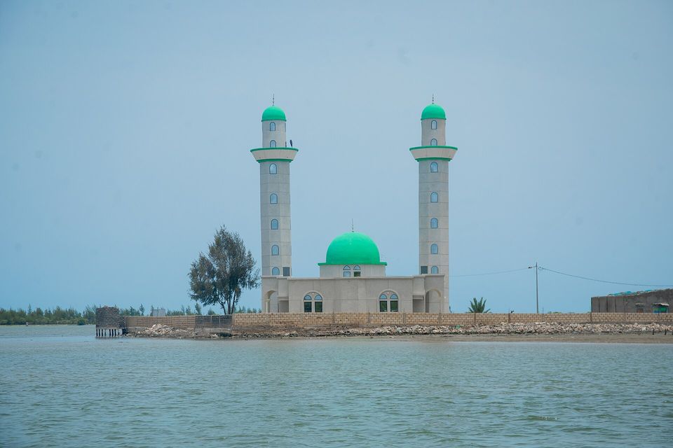 A white mosque with a green dome and two tall green-capped minarets stands on a shell-walled shore beside a single tree, calm grey-green water in front